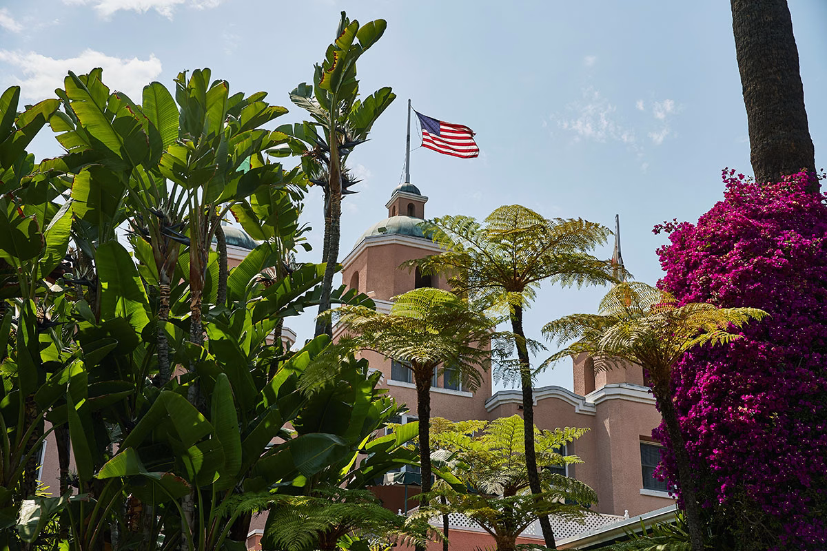 Die rosafarbenen Türme des Hauptgebäudes des The Beverly Hills Hotel mit einer wehenden US-Flagge auf der Spitze, eingerahmt von tropischen Palmen, großen Farnbäumen und leuchtend lila Bougainvillea-Blüten