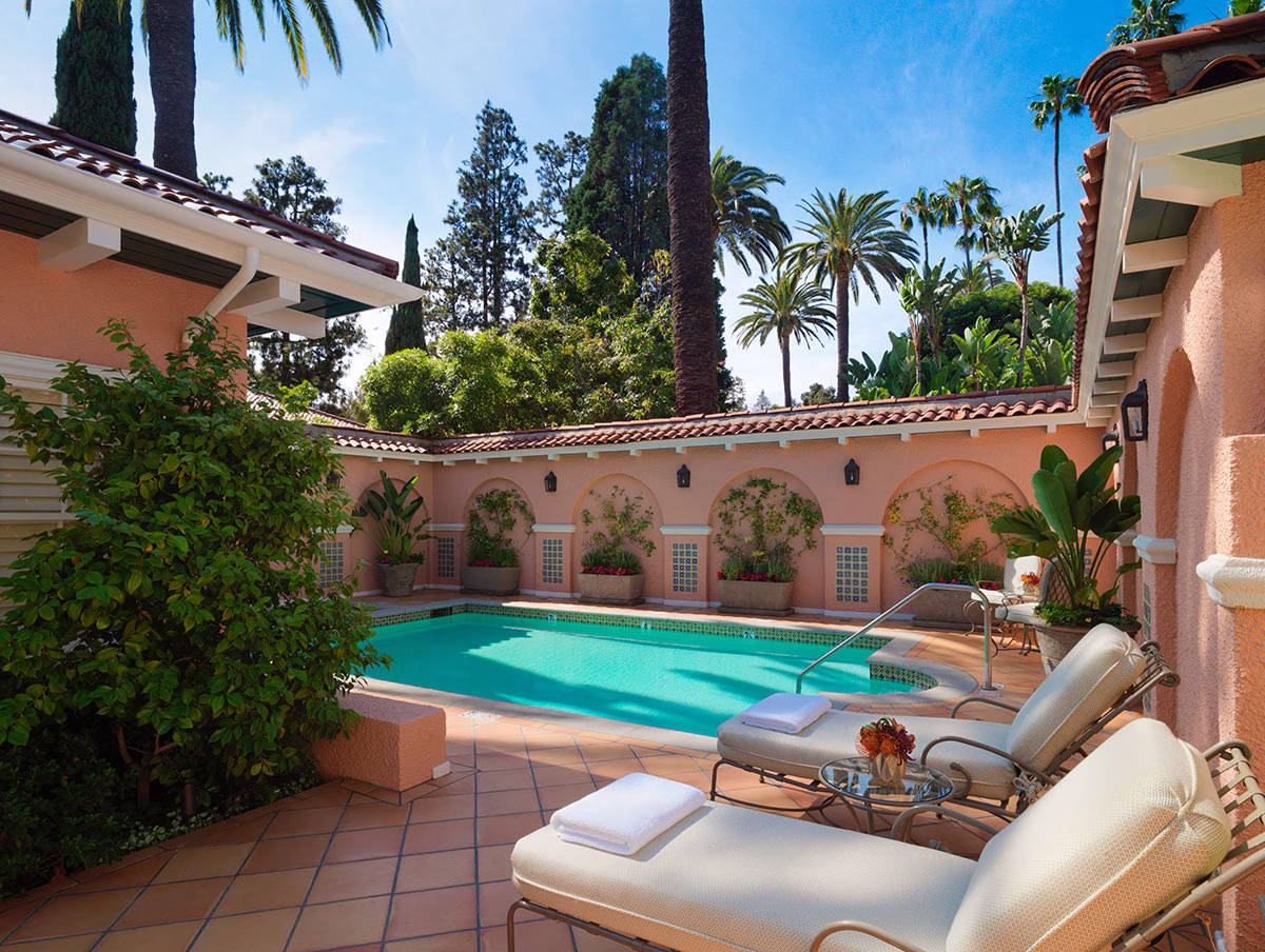 Der private Innenhof von Bungalow 5A mit einem türkisfarbenen Swimmingpool, umgeben von einer rosafarbenen Mauer mit Rundbögen, hellen Liegestühlen auf Terrakotta-Fliesen und hohen Palmen im Hintergrund