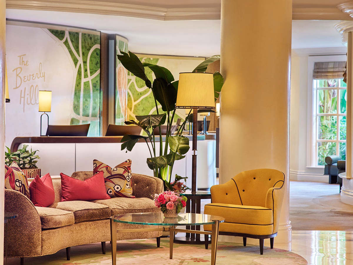 Die stilvolle Hotel-Lobby mit einer geschwungenen Rezeption im Hintergrund, einer gemütlichen beigen Couch mit roten Kissen, einem gelben Sessel im Retro-Stil und einer großen Zimmerpalme neben einer Stehlampe