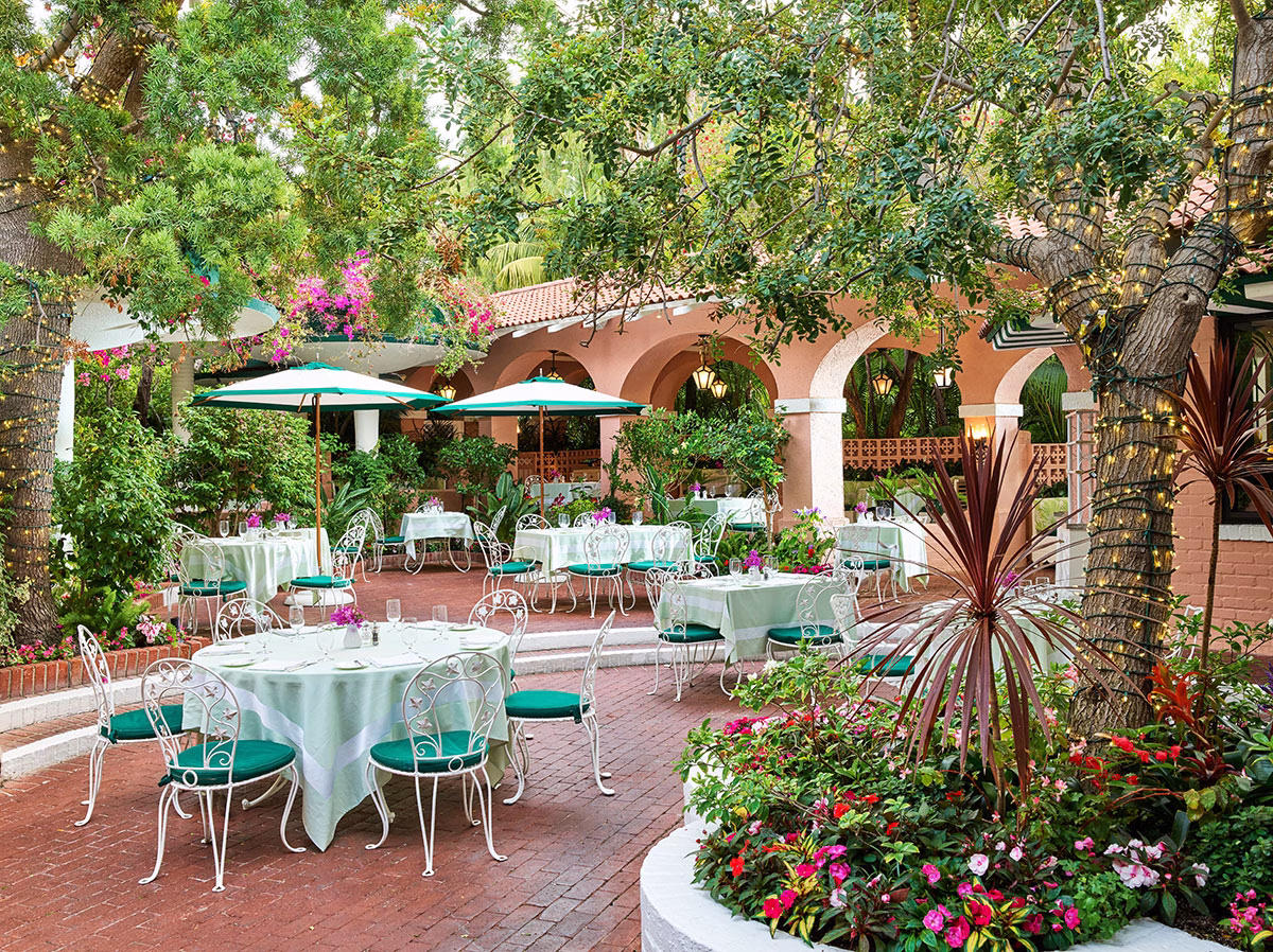 Die idyllische Außenterrasse der Polo Lounge mit rot gepflastertem Boden, weiß-grünen Sonnenschirmen über eleganten Tischen, umgeben von mit Lichterketten geschmückten Bäumen und üppigen Blumenbeeten vor rosafarbenen Arkadenbögen