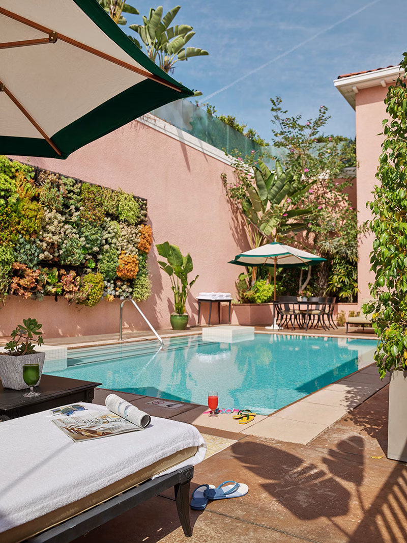 Der private Außenpoolbereich des Presidential Bungalow 24A mit einem türkisfarbenen Pool, einer üppig bepflanzten vertikalen Gartenwand an einer rosafarbenen Mauer, einer gemütlichen Sonnenliege mit Magazin im Vordergrund und Sitzgelegenheiten unter grün-weißen Schirmen