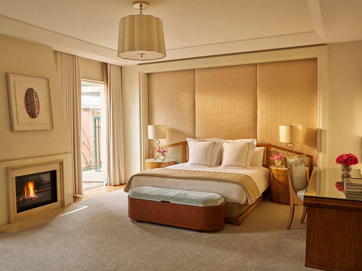 Das luxuriöse Hauptschlafzimmer des Presidential Bungalow 24A mit einem eleganten Kingsize-Bett vor einer cremefarbenen, gepolsterten Wand, einem brennenden Kamin auf der linken Seite, einer stilvollen Sitzecke und einem edlen Holzschreibtisch im Vordergrund