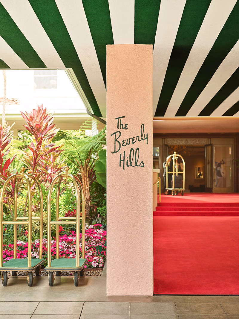 Nahaufnahme einer rosafarbenen Säule mit dem eleganten, dunkelgrünen Schriftzug „The Beverly Hills“ am Hoteleingang, neben dem zwei goldene Gepäckwagen auf dem roten Teppich vor einer blühenden Blumenrabatte stehen