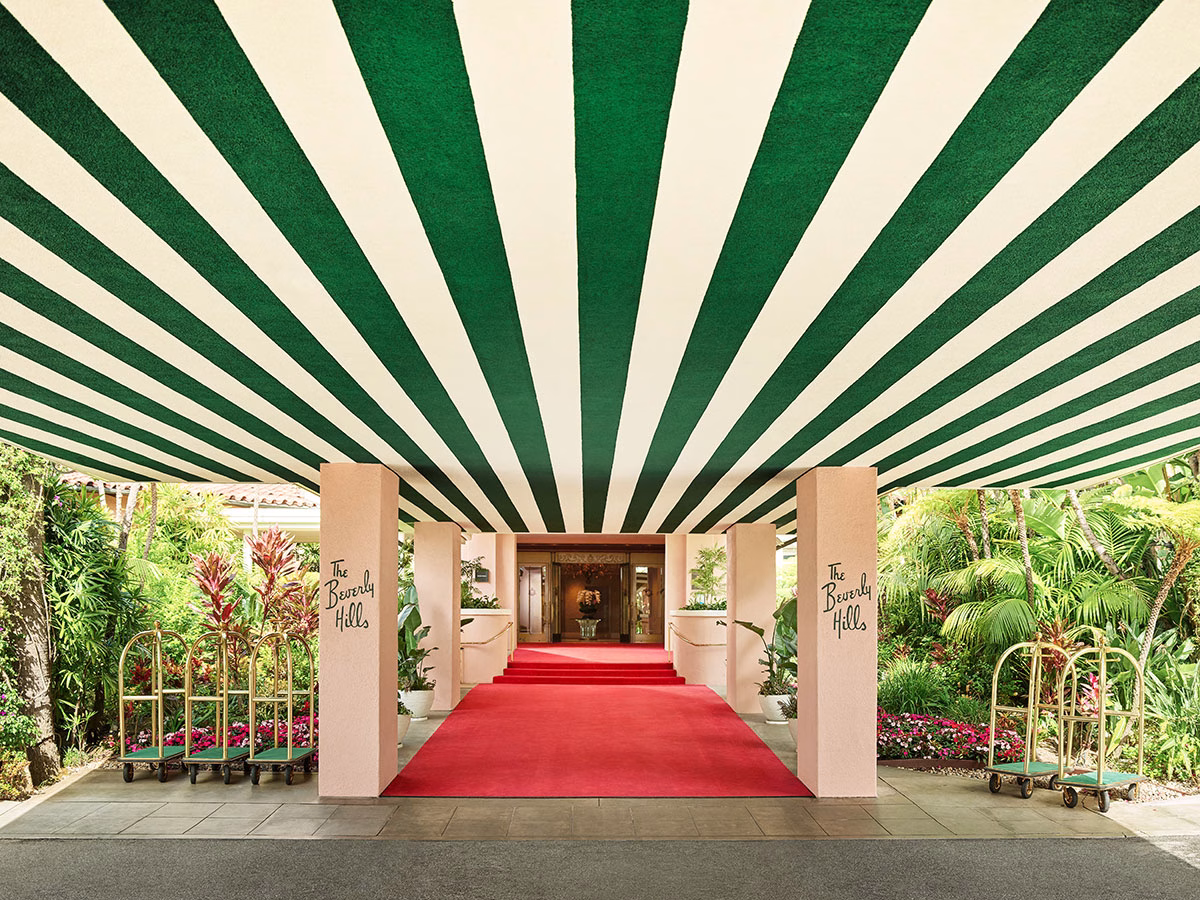 Der berühmte Haupteingang des The Beverly Hills Hotel mit einem breiten roten Teppich, der unter einer markanten grün-weiß gestreiften Überdachung hindurchführt, flankiert von hellrosa Säulen mit dem geschwungenen Hotel-Schriftzug und goldenen Gepäckwagen