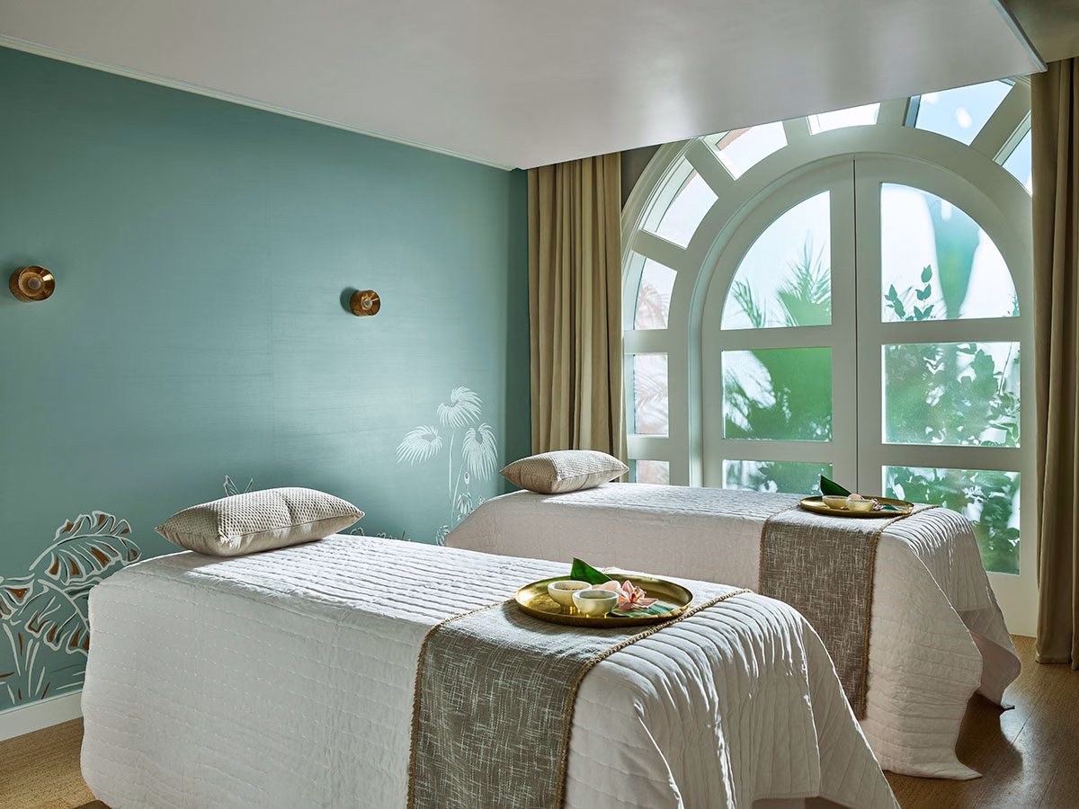 Die stilvolle Spa-Paarsuite mit zwei weiß gedeckten Massageliegen, auf denen goldene Tabletts mit Schalen bereitstehen, vor einer beruhigenden, salbeigrünen Wand mit dezenten Palmen-Illustrationen und einem großen Rundbogenfenster