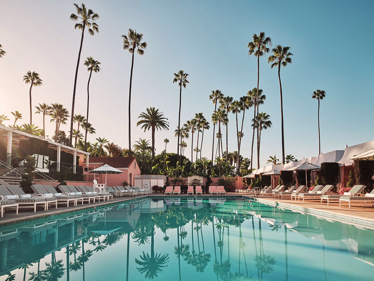 Der große, von Palmen umgebene Swimmingpool des The Beverly Hills Hotel, in dessen klarem Wasser sich die riesigen Palmen spiegeln, flankiert von grün-weiß gestreiften und rosafarbenen Liegestühlen sowie weißen Cabanas unter blauem Himmel