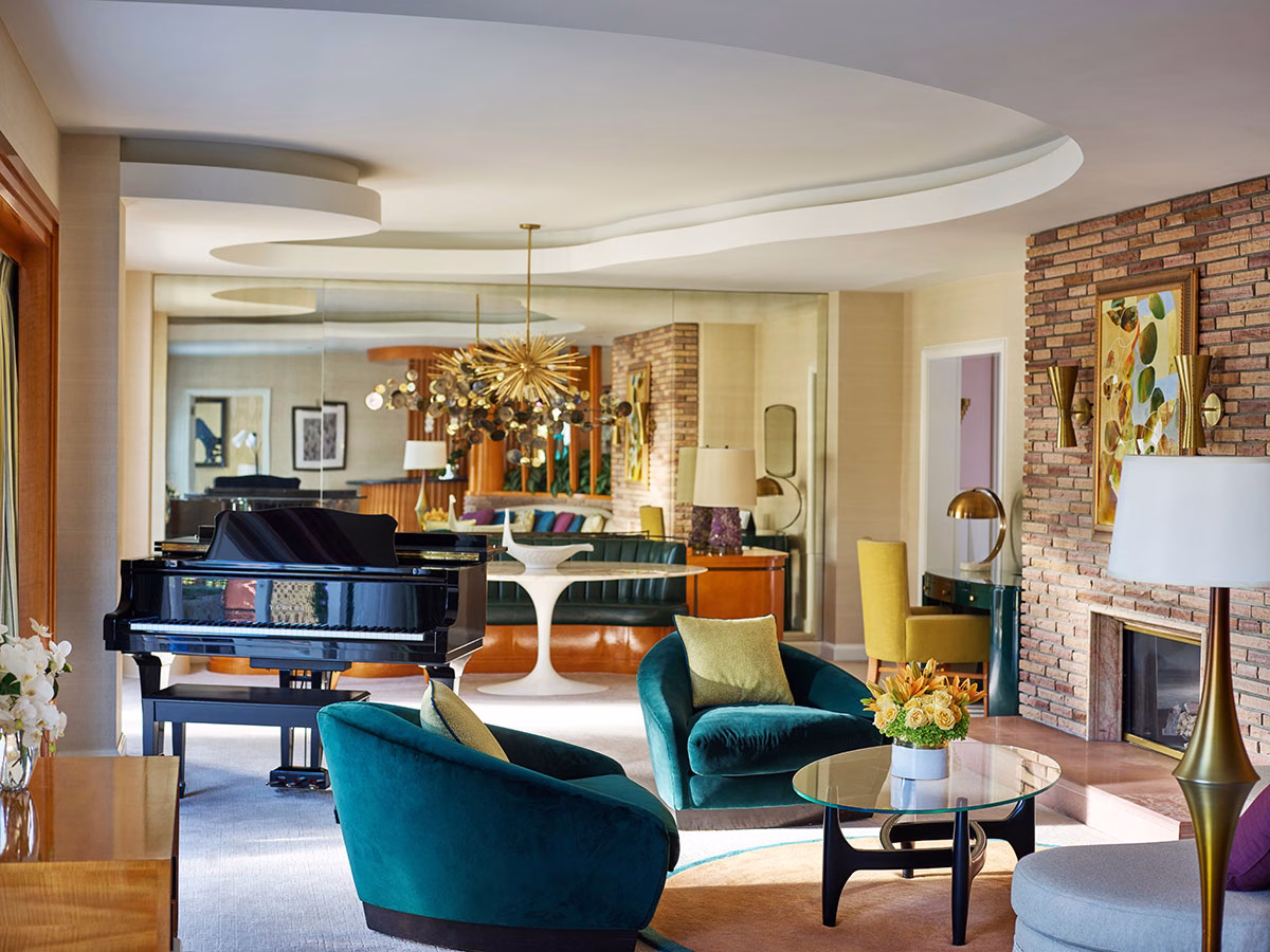 Das elegante Wohnzimmer der Paul R. Williams Suite mit einem glänzenden schwarzen Konzertflügel, zwei tiefgrünen Samtsesseln um einen Glastisch und einer gemütlichen Kaminecke vor einer rustikalen Backsteinwand