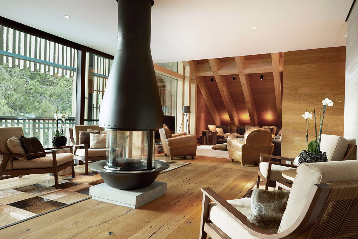Großzügiger Wohnbereich der Deluxe-Suite im The Chedi Andermatt mit edlem Holzfußboden, gemütlichen Sesseln und einem freistehenden, modernen Kamin aus schwarzem Metall im Zentrum des Raumes