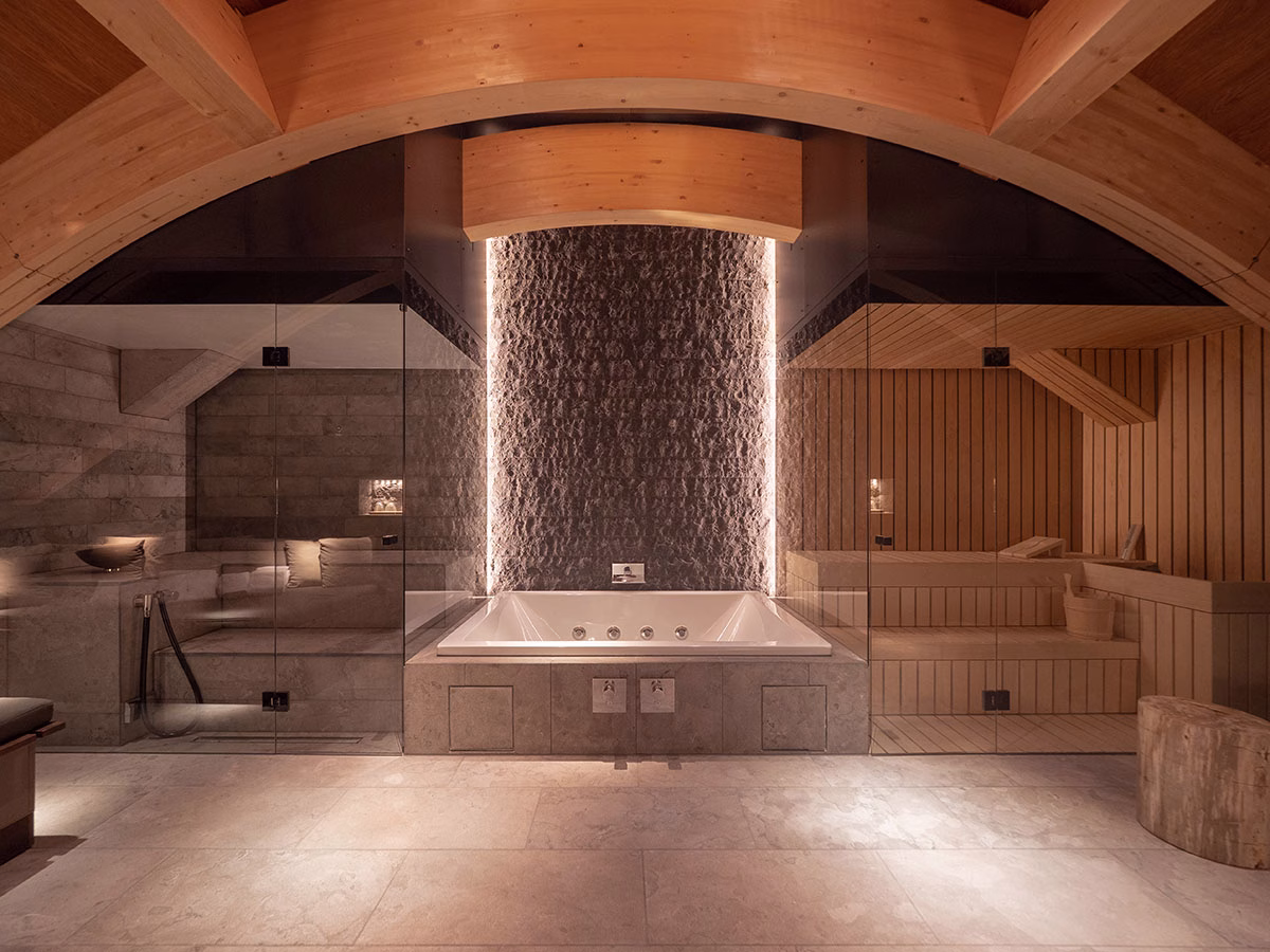 Exklusiver Spa-Bereich im The Chedi Andermatt unter einer hölzernen Dachschräge, bestehend aus einem eingelassenen Whirlpool, flankiert von einer verglasten Sauna auf der rechten und einem Dampfbad auf der linken Seite