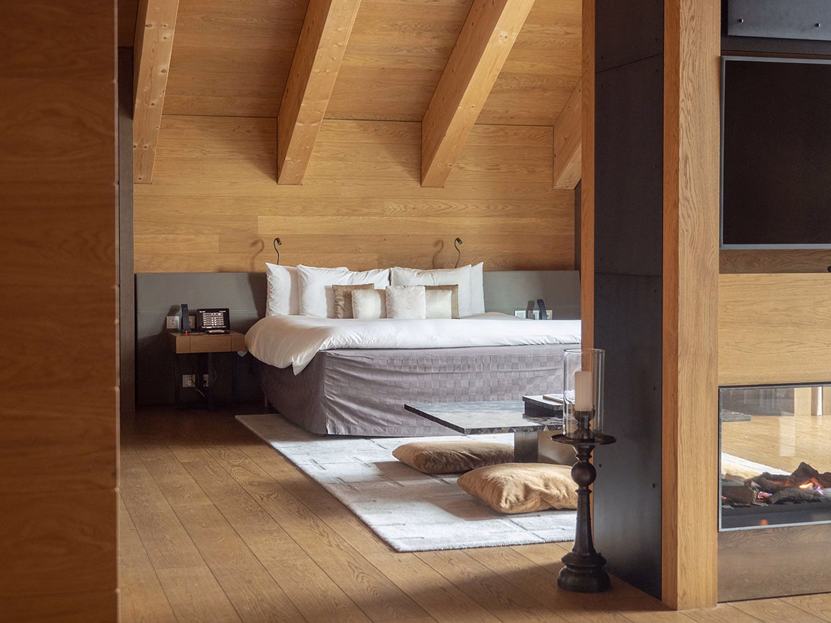 Ein stilvolles Schlafzimmer in der Furka-Suite des The Chedi Andermatt mit einem großen Doppelbett unter Sichtbalken aus Holz, einer gemütlichen Sitzecke und einem modernen Glaskamin im Vordergrund