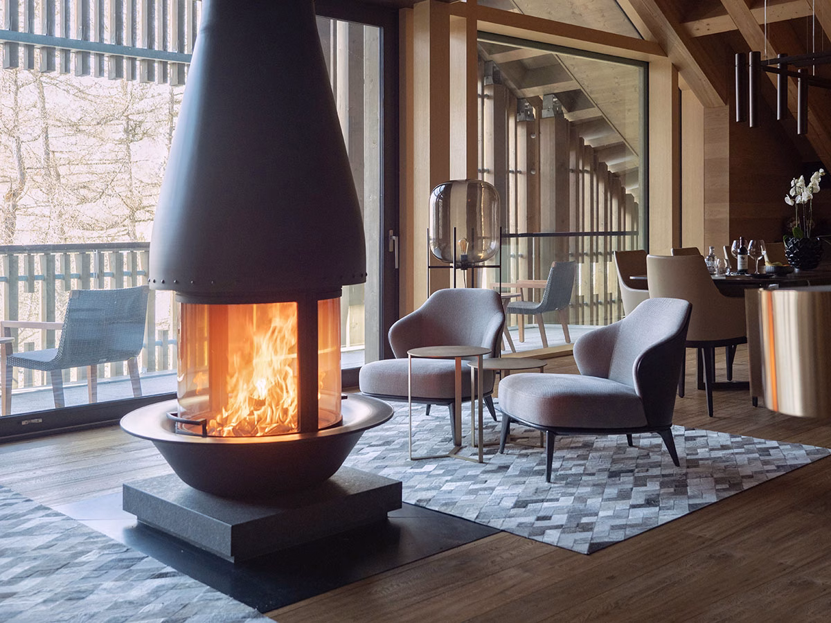 Der luxuriöse Wohn- und Essbereich der Gotthard-Suite im The Chedi Andermatt mit einem prasselnden, offenen Kamin aus schwarzem Metall, grauen Design-Sesseln und einer großen Fensterfront zum Balkon