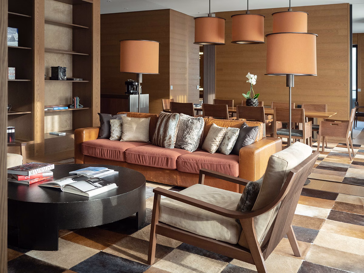 Luxuriöser Wohnbereich der Grand Deluxe Suite im The Chedi Andermatt mit einem cognacfarbenen Ledersofa, gemustertem Teppich, einem runden Couchtisch und einem langen Esstisch aus Holz im Hintergrund