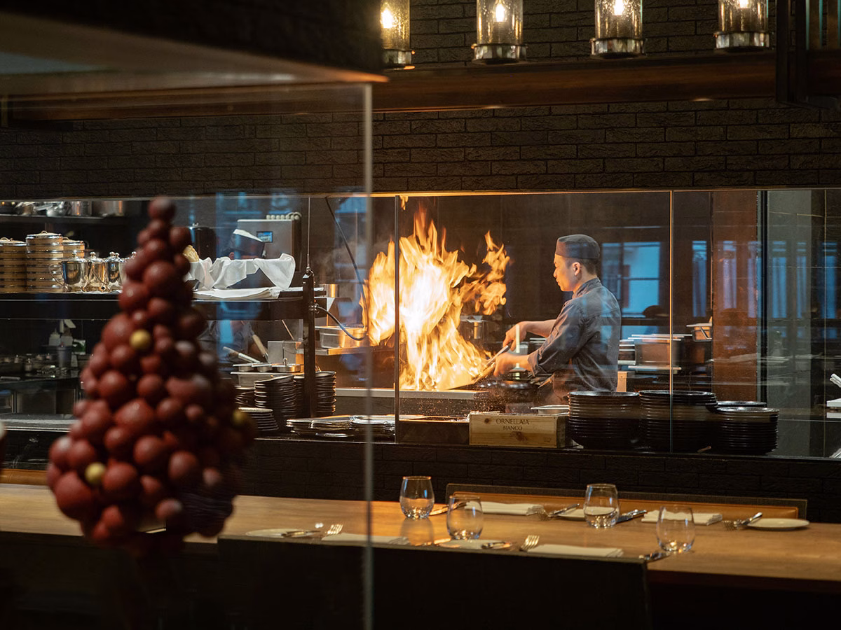 Blick durch eine Glasscheibe in die asiatische Showküche des The Chedi Andermatt, in der ein Koch in grauer Uniform an einem züngelnden Wok-Feuer arbeitet