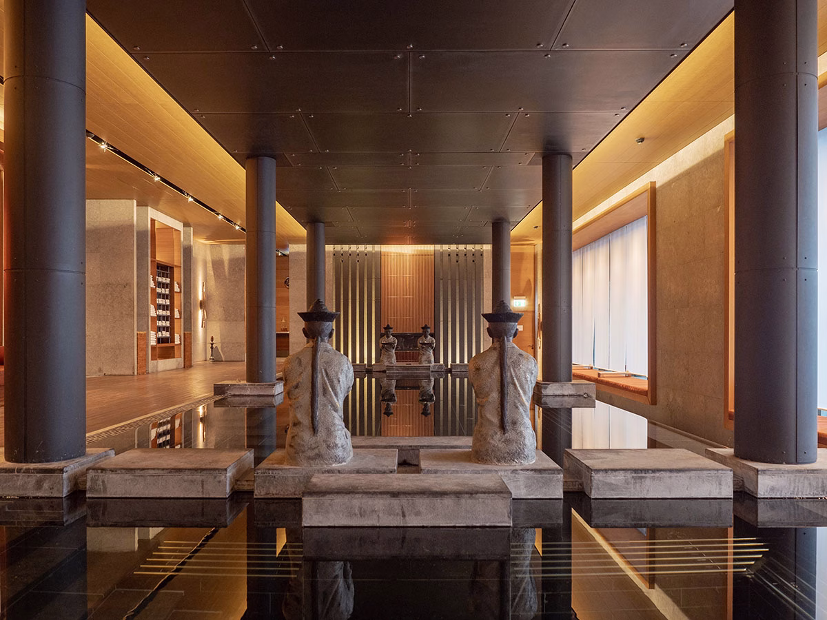 Symmetrisch gestaltetes Tempel-Bassin im Spa des The Chedi Andermatt mit dunklen Säulen und steinernen asiatischen Wächterfiguren, deren Silhouetten sich im spiegelglatten Wasser reflektieren