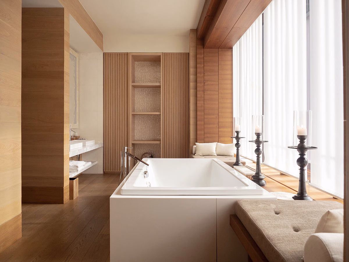 Ein elegantes Badezimmer im The Chedi Andermatt mit einer freistehenden, weißen Badewanne vor einer bodentiefen Fensterfront mit weißen Vorhängen und großen, schwarzen Kerzenleuchtern