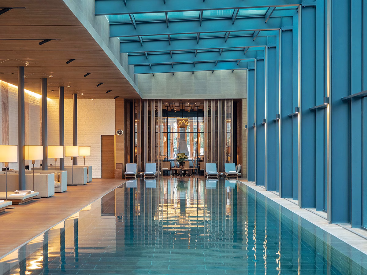 Der monumentale Indoor-Pool im The Chedi Andermatt unter einer verglasten Deckenkonstruktion, gesäumt von beigen Lounge-Betten auf der linken Seite und einer blau schimmernden Fensterfront auf der rechten Seite