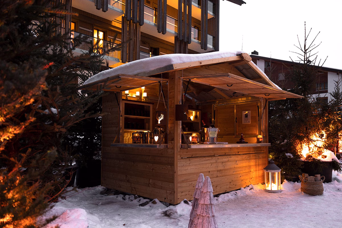 Eine urige, schneebedeckte Holzhütte auf dem Außengelände des The Chedi Andermatt, die am Abend beleuchtet als winterliche Bar im Freien dient, umgeben von Tannenbäumen