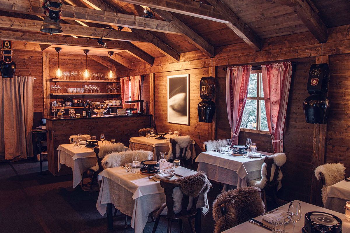 Das gemütliche Innere des Restaurants „The Chalet“ im The Chedi Andermatt, rustikal eingerichtet mit Holzwänden, Schweizer Kuhglocken, Vorhängen im rot-weißen Karomuster und mit Fell ausgelegten Stühlen