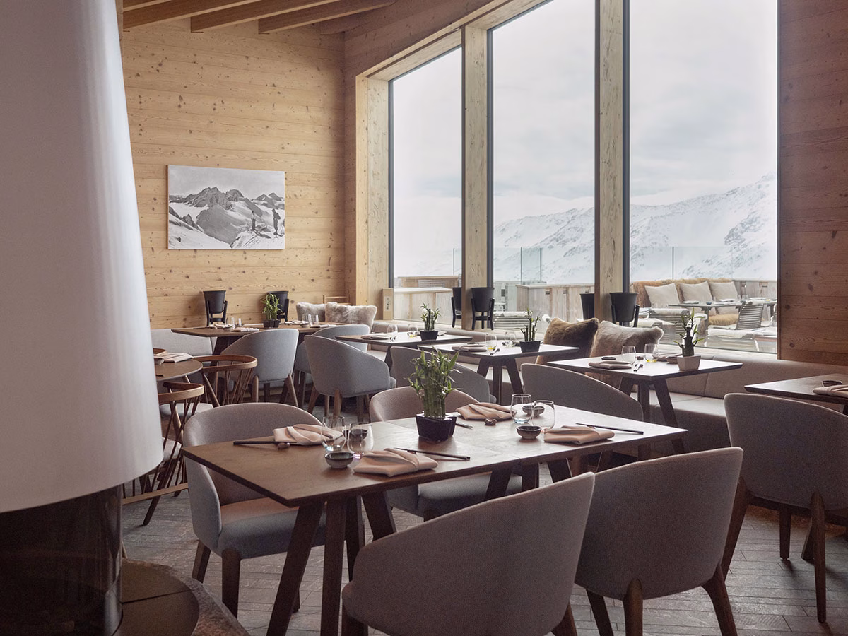 Hell erleuchteter Restaurantbereich im The Chedi Andermatt mit modernen, grau polsterten Sesseln an dunklen Holztischen vor einer riesigen Panoramafensterfront, die den Blick auf die verschneiten Schweizer Alpen freigibt