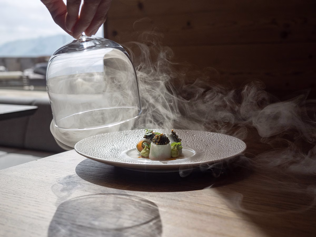 Close-up einer Hand, die eine gläserne Servierglocke von einem kunstvoll angerichteten Kaviar-Gericht hebt, während dichter, weißer Rauch effektvoll über den hölzernen Restauranttisch strömt