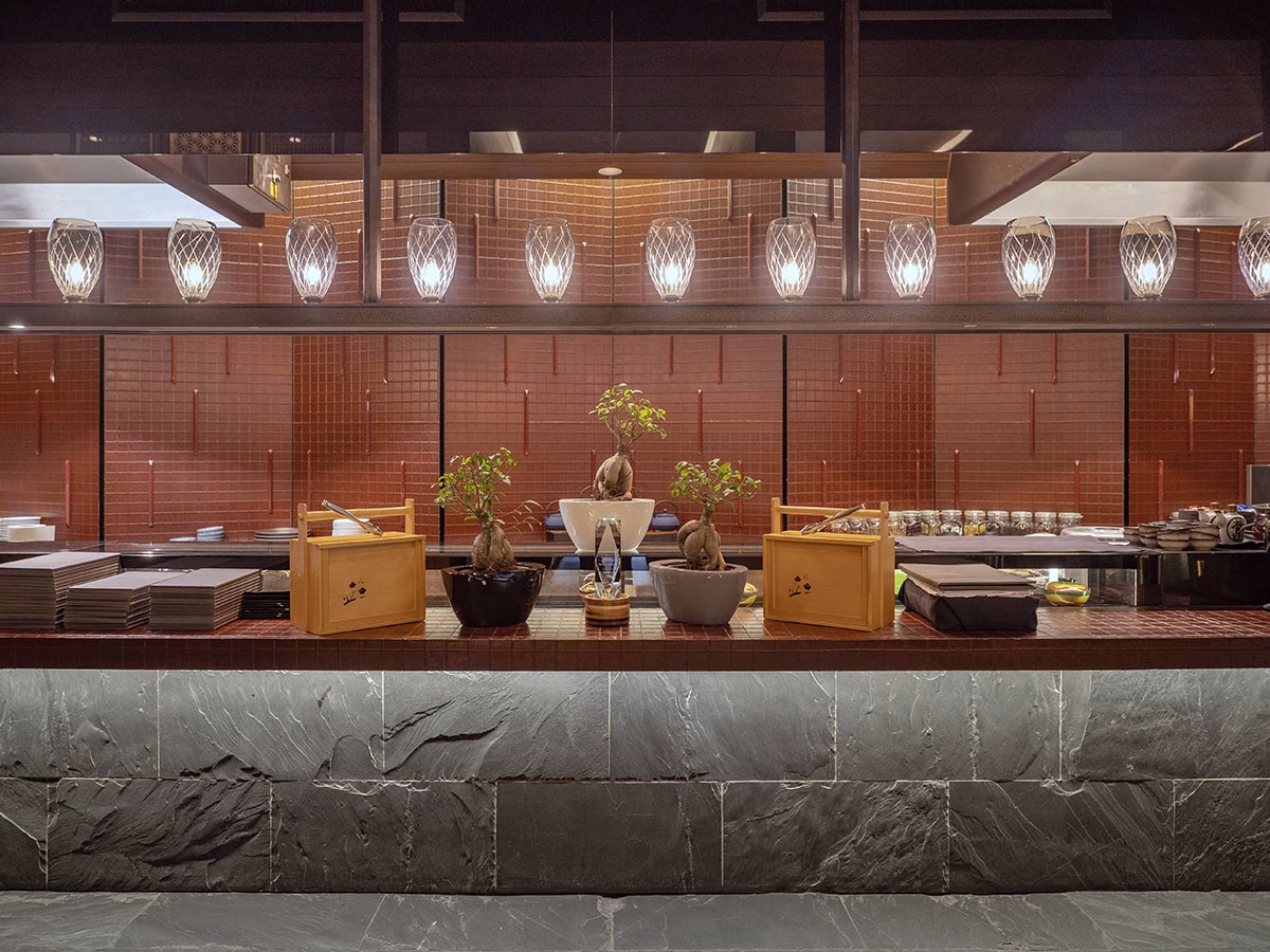 Der edle Tresen im „The Japanese Restaurant“ des The Chedi Andermatt, gestaltet mit einer dunklen Schieferfront, kleinen Bonsai-Bäumchen zur Dekoration und hängenden Glasleuchten vor einer rotbraun gefliesten Wand