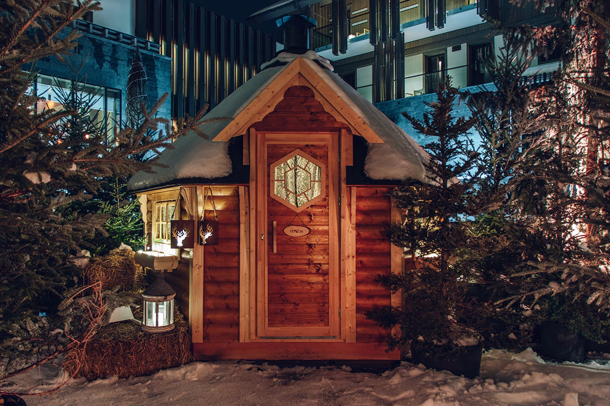 Eine kleine, urige Holzhütte mit schneebedecktem Dach auf dem winterlichen Außengelände des The Chedi Andermatt bei Nacht, stimmungsvoll beleuchtet und umgeben von Tannenbäumen und Strohballen