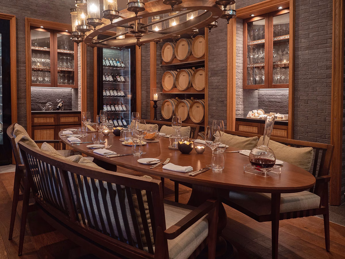 Der exklusive Wein- und Käsekeller des The Chedi Andermatt mit einem großen, ovalen Holzesstisch, umgeben von Weinregalen, aufgereihten Holzfässern und einer Wand aus dunklem Backstein unter einem rustikalen Kronleuchter