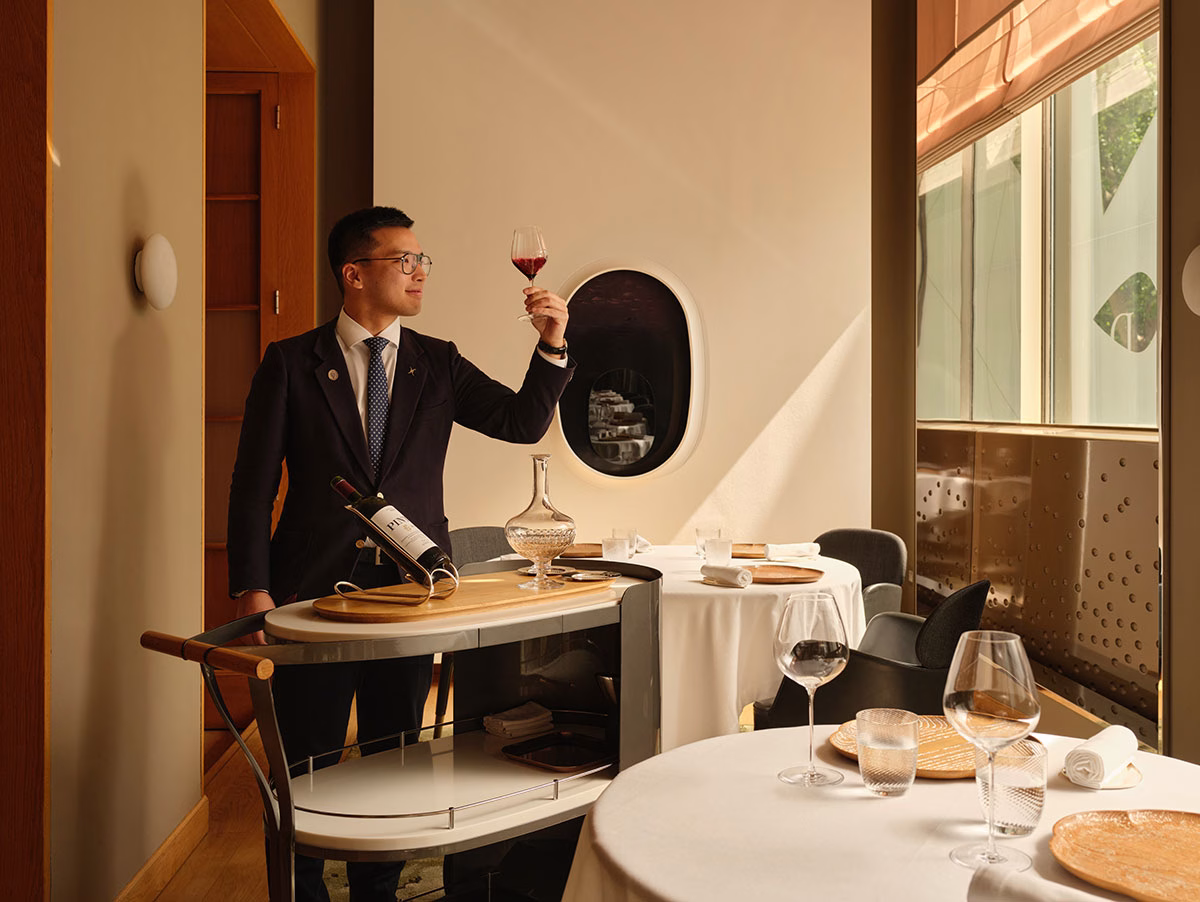 Ein eleganter Sommelier im feinen Anzug begutachtet ein Glas Rotwein im sternengekrönten Restaurant Alain Ducasse at The Dorchester, neben einem modernen, stilvollen Weinservierwagen