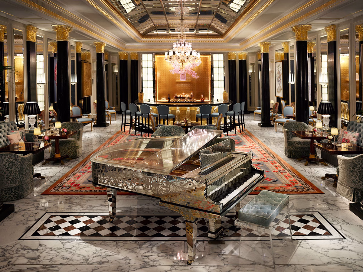 Die opulente Promenade-Lobby im Hotel The Dorchester mit prachtvollen Marmorsäulen, einem glänzenden, verspiegelten Konzertflügel im Vordergrund und einer golden schimmernden Bar im Hintergrund