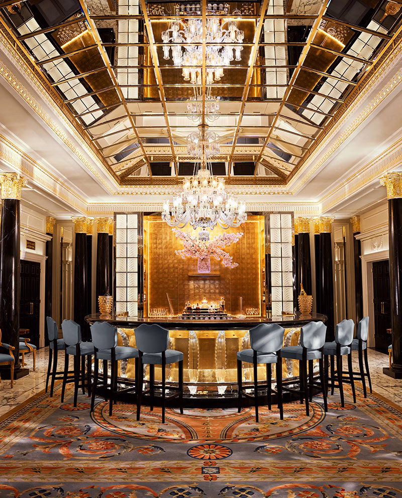 Der prunkvolle Barbereich im Hotel The Dorchester mit einer geschwungenen, goldenen Bartheke, blaugrauen Samt-Barhockern und einer riesigen, spiegelnden Glasdecke, die das warme Licht reflektiert