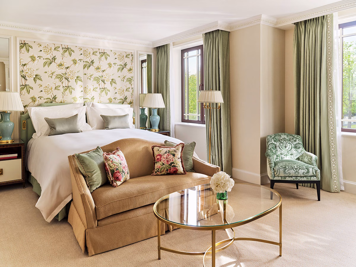Ein britisches Luxushotelzimmer im The Dorchester mit einer floral gemusterten Tapete, einem Kingsize-Bett, salbeigrünen Vorhängen und einem eleganten ockerfarbenen Sofa am Fußende