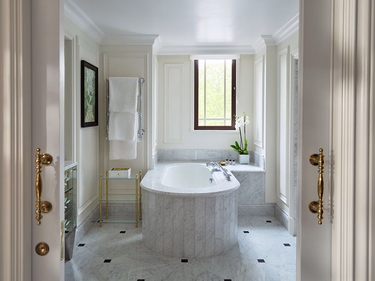 Blick durch weiße Flügeltüren mit goldenen Beschlägen in ein helles Badezimmer im The Dorchester mit einer ovalen, weiß verkleideten Marmorbadewanne unter einem Fenster