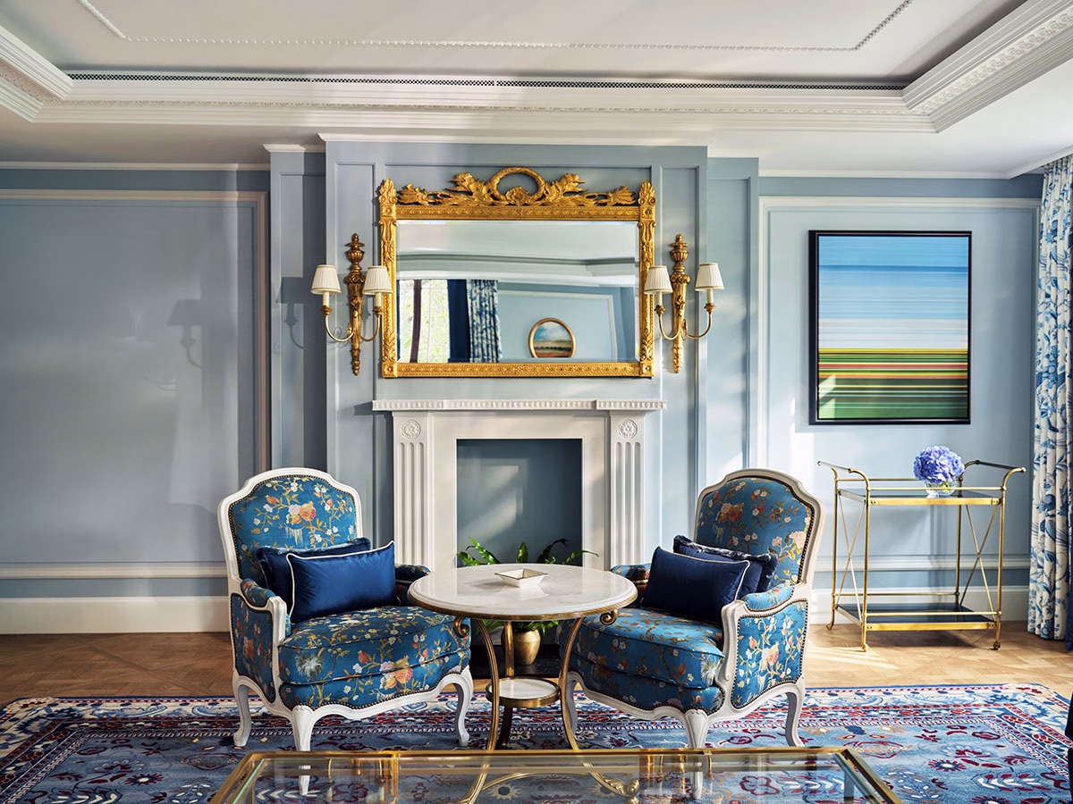 Ein elegant gestalteter Salon in Pastellblau im Hotel The Dorchester mit zwei floral gemusterten Sesseln, einem edlen weißen Kamin und einem großen Prunkspiegel mit goldenem Barockrahmen