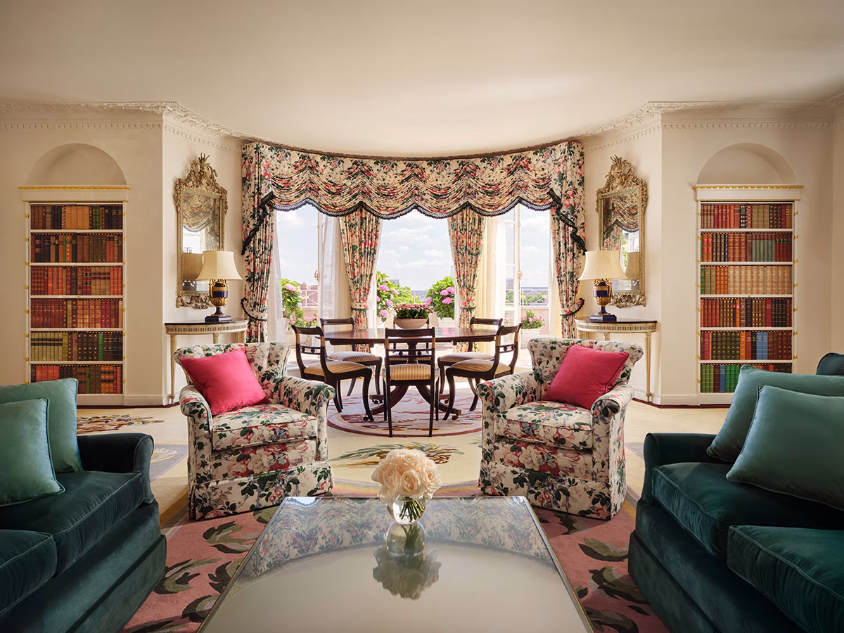 Ein herrschaftliches Wohnzimmer im englischen Stil mit floralen Mustern, samtigen dunkelgrünen Sofas, zwei Einbaubücherregalen voller klassischer Bände und einer großen Fensterfront mit Blick auf die Terrasse