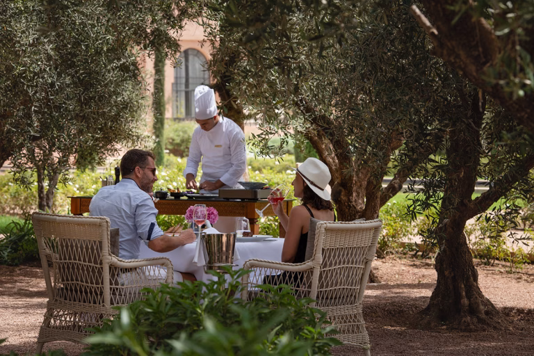 Ein Pärchen sitzt an einem Tisch und wird zwischen Bäumen im Garten des The Oberoi Marrakech von einem privaten Chef bekocht