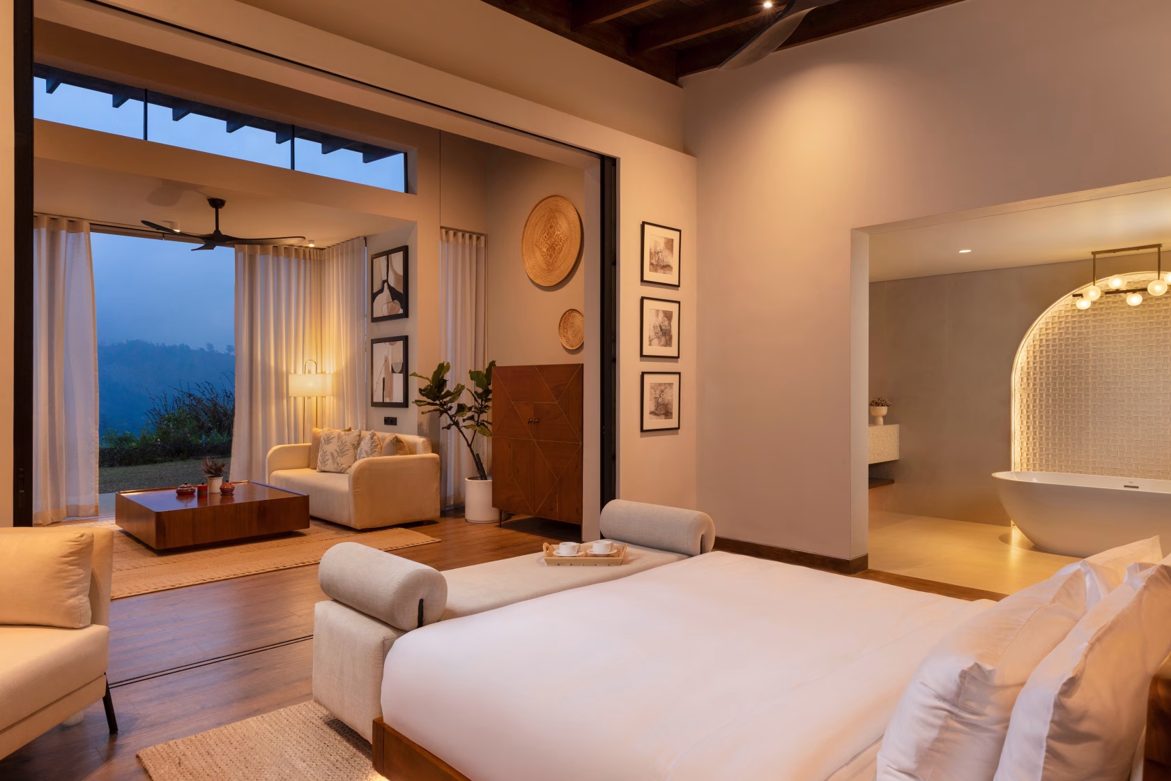 Blick aus dem Schlafbereich einer Suite im Uga Ghiri über das elegant gemachte Doppelbett hinweg auf den angrenzenden Wohnbereich mit Sofa und das offene, hell erleuchtete Badezimmer mit Badewanne