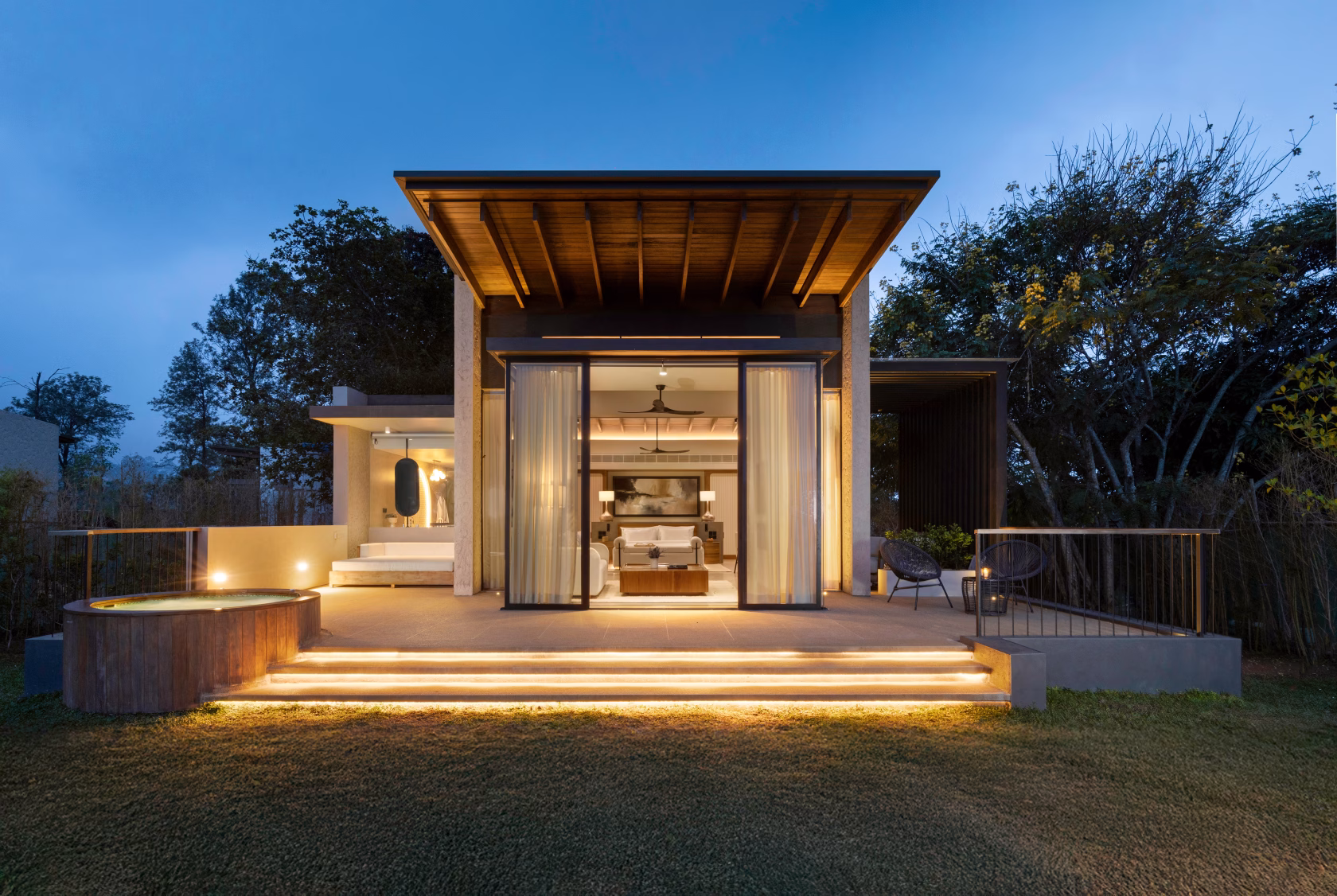 Außenansicht einer luxuriösen Villa im Uga Ghiri bei Dämmerung mit beleuchteten Stufen, einer Terrasse samt rundem Whirlpool und weit geöffneten Glasschiebetüren, die den Blick auf das elegante Schlafzimmer freigeben