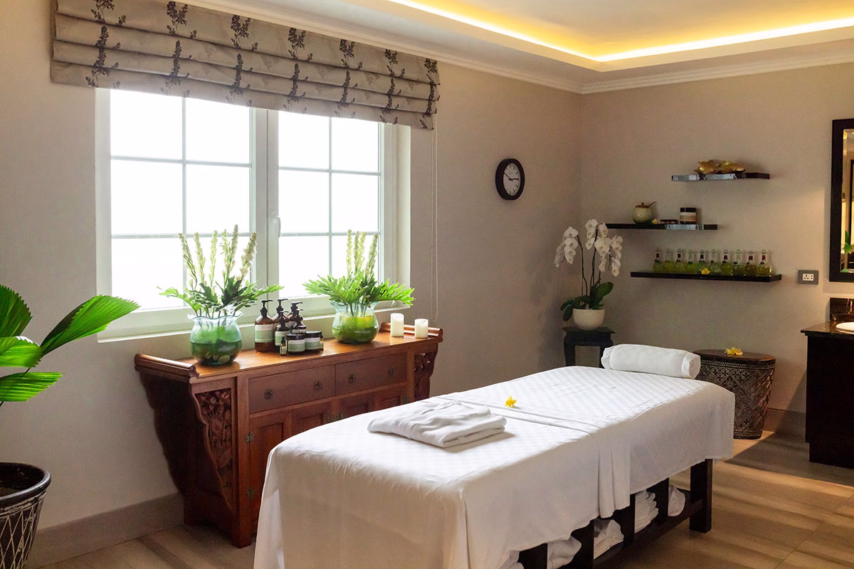 Der helle Behandlungsraum im Akoya Spa des Viceroy Bali mit einer weiß gedeckten Massageliege, einem eleganten Holz-Sideboard am großen Fenster und frischen Orchideen vor einer cremefarbenen Wand