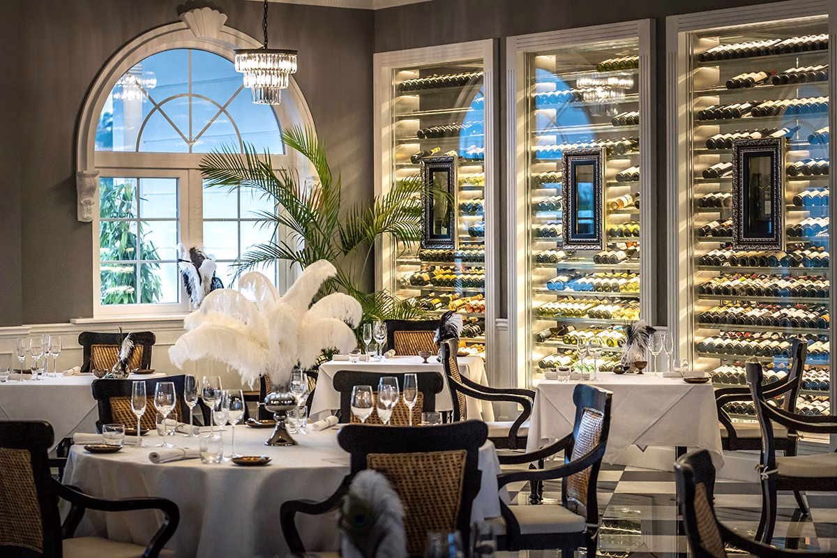 Das stilvolle Interieur des Apéritif Restaurants im Viceroy Bali im edlen Art-déco-Design mit weiß eingedeckten Tischen, großen, beleuchteten Weinschränken im Hintergrund und eleganter Feder-Dekoration