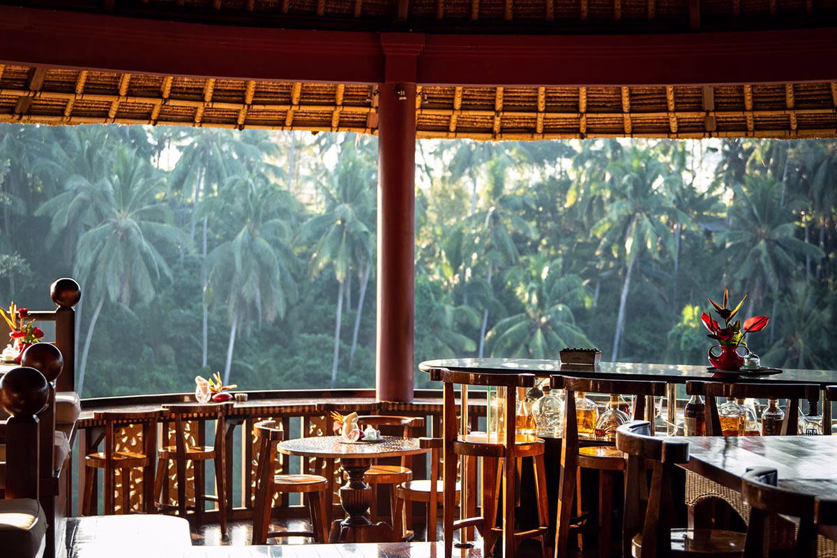 Blick aus der gemütlichen Bar des Cascades Restaurants im Viceroy Bali mit Holzstühlen und -tischen unter einem traditionellen Reetdach auf das dicht bewachsene, tropische Dschungeltal von Ubud