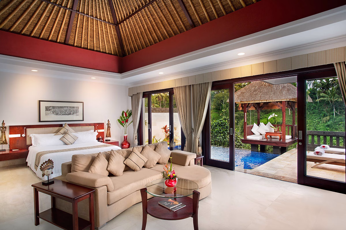 Innenansicht einer Deluxe Terrace Villa im Viceroy Bali mit einem großen Kingsize-Bett unter einer hohen Holzdecke, einer beigen Sofagarnitur und geöffneten Glasschiebetüren, die direkt zum privaten Infinity-Pool und einer traditionellen Bale führen