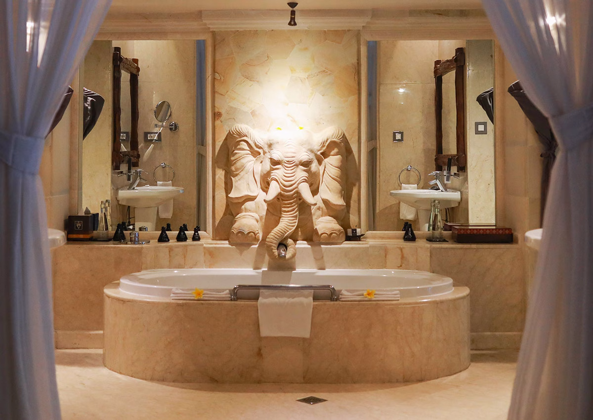 Das luxuriöse Badezimmer der Elephant Villa im Viceroy Bali mit einer großen, ovalen Marmor-Badewanne, die von einer kunstvollen, direkt in die Steinwand gemeißelten Elefantenskulptur verziert wird