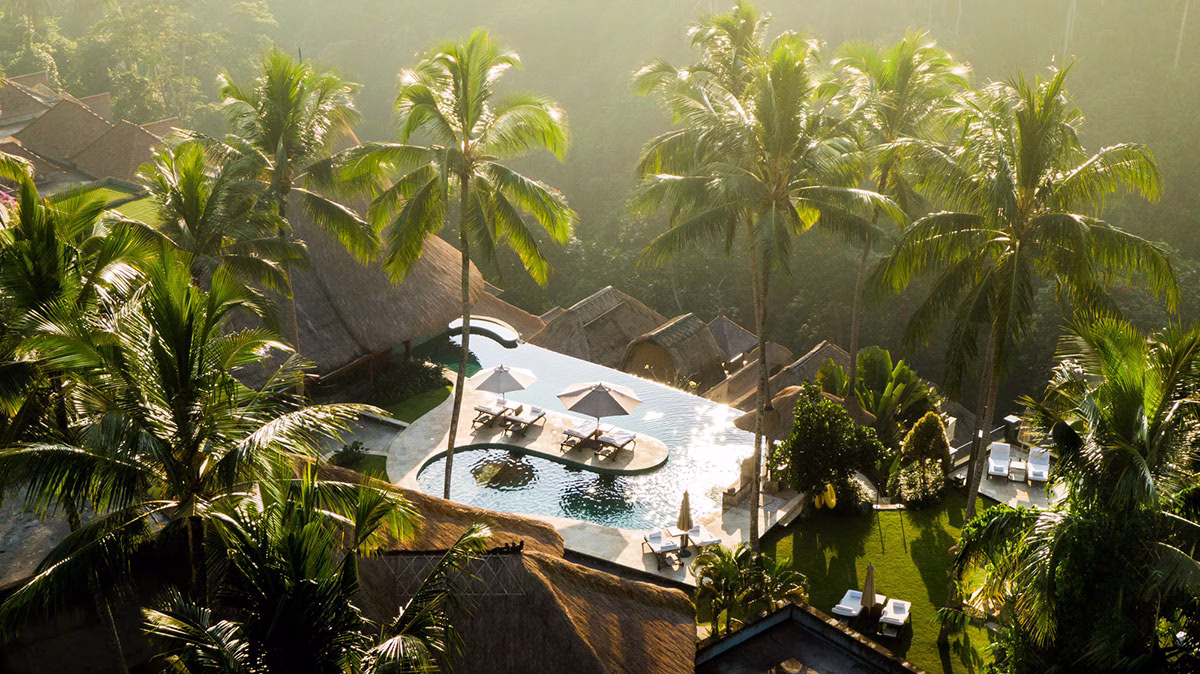 Drohnen-Aufnahme aus der Vogelperspektive auf den Infinity-Hauptpool des Viceroy Bali, der von hohen Palmen, Sonnenliegen mit Schirmen und den traditionellen Dächern des Luxusresorts umgeben im Sonnenlicht glänzt
