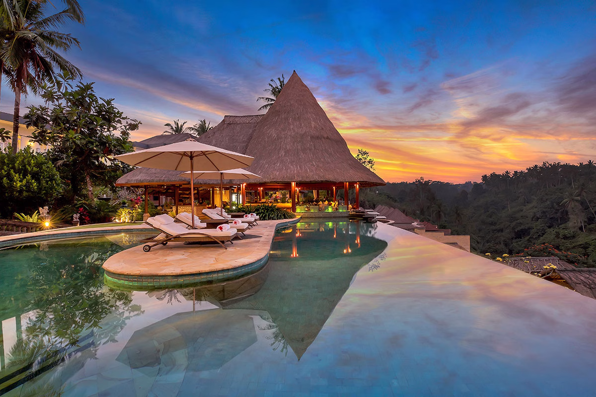 Der Infinity-Hauptpool des Viceroy Bali bei einem spektakulären Sonnenuntergang, dessen farbenprächtiger Abendhimmel sich im ruhigen Poolwasser spiegelt, während die Liegen und das reetgedeckte Hauptgebäude beleuchtet sind