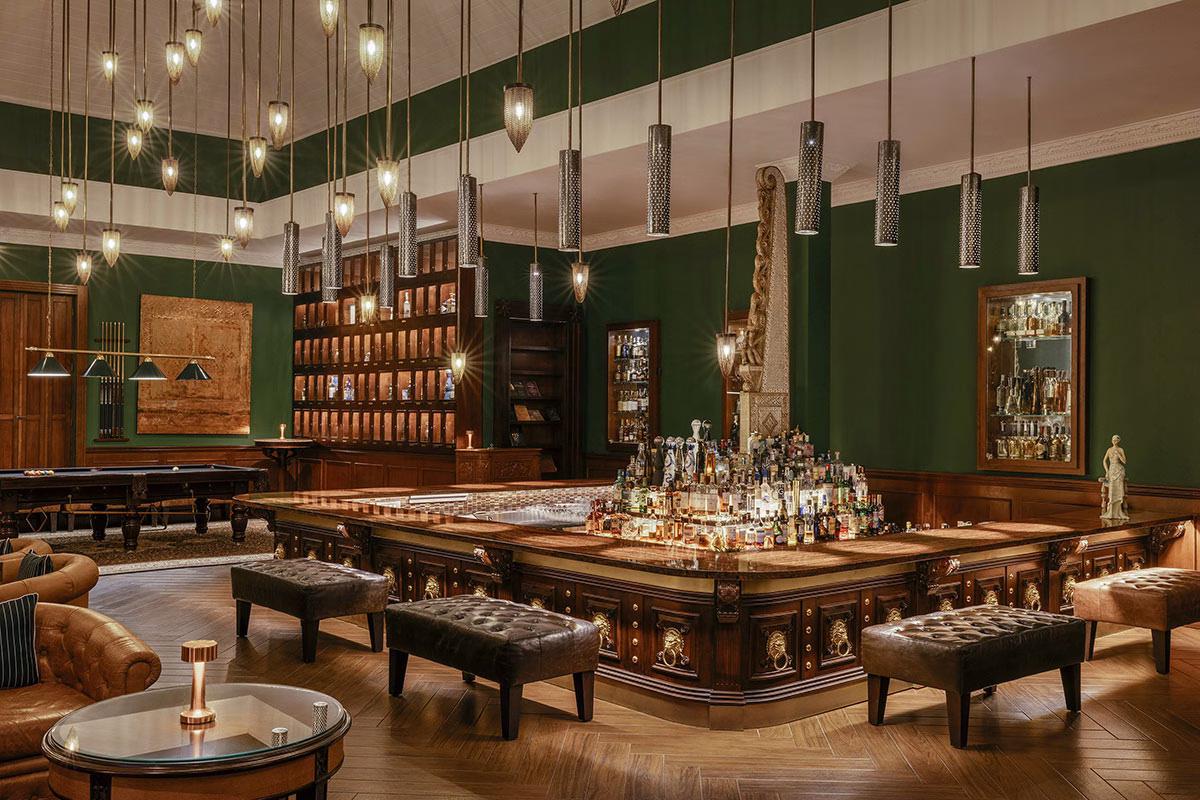 Das luxuriöse Interieur der Pinstripe Bar im Viceroy Bali mit dunkelgrünen Wänden, einer edlen, quadratischen Holzbar mit beleuchteter Spirituosen-Auswahl in der Mitte, Chesterfield-Sesseln und zahlreichen von der Decke hängenden Designer-Leuchten