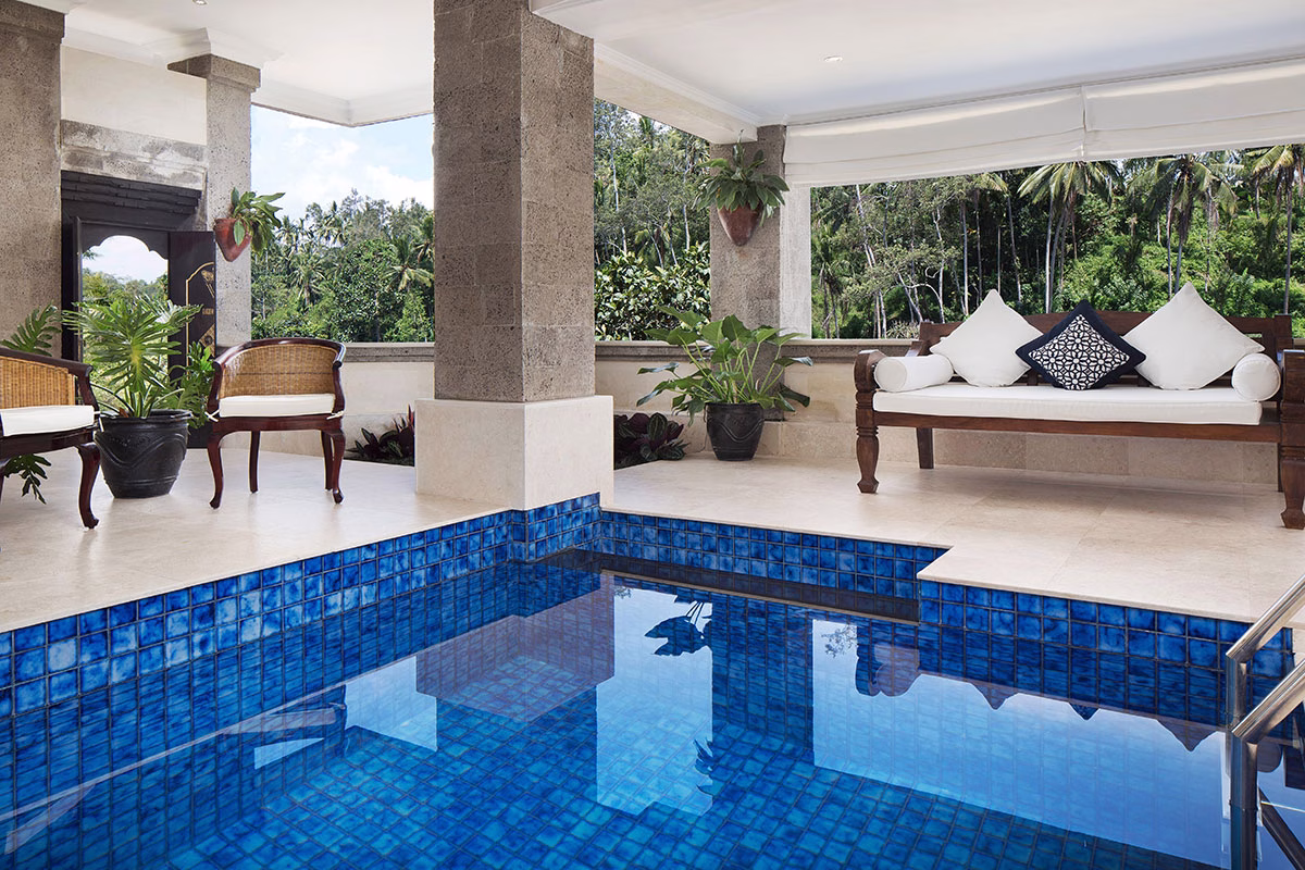 Ein privater, überdachter Loungebereich einer Pool Suite im Viceroy Bali mit einem blauen Tauchbecken im Vordergrund, beigen Korbsesseln und einem eleganten weißen Daybed vor einer Kulisse aus Palmen
