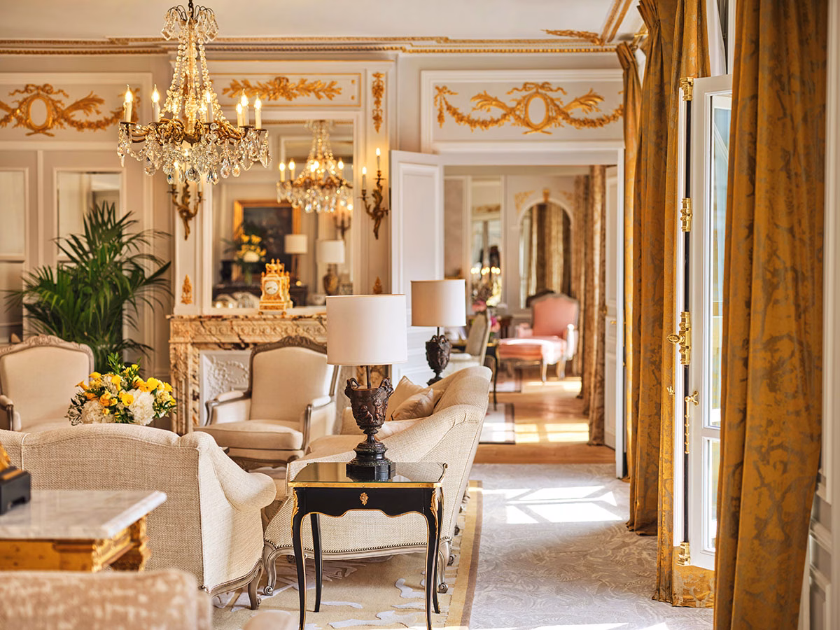 Das luxuriöse Wohnzimmer der Royale Suite 509 im Hôtel Plaza Athénée mit vergoldeten Wandverzierungen, einem Kristallkronleuchter, eleganten beigen Sofas, goldgelben Vorhängen und einem Durchblick in ein rosa gestaltetes Nebenzimmer