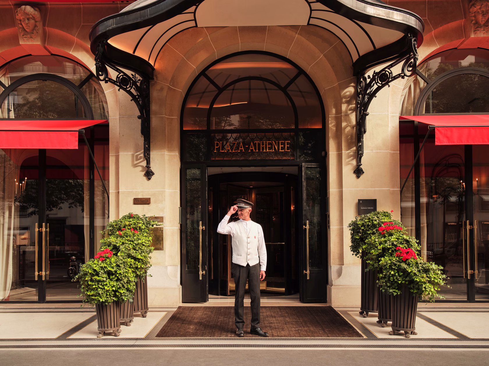 Ein Portier in weißer Uniform und Schirmmütze steht grüßend unter dem eleganten, gläsernen Vordach des Haupteingangs des Hôtel Plaza Athénée, flankiert von roten Markisen und großen, grünen Pflanzkübeln mit roten Geranien