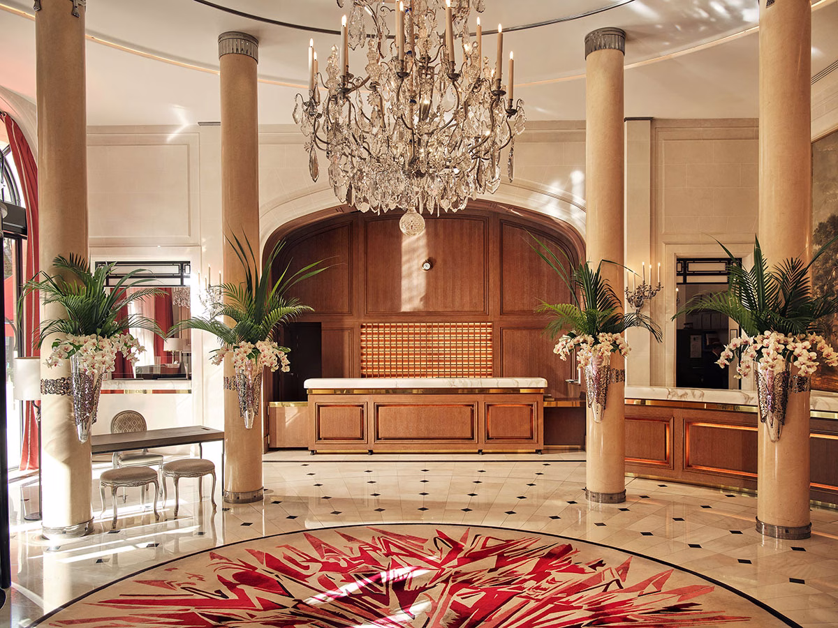 Die luxuriöse Hotel-Lobby des Hôtel Plaza Athénée mit einem prachtvollen, zentralen Kristallkronleuchter, eleganten sandfarbenen Säulen, kunstvollen Blumengestecken aus Orchideen und einem auffälligen Teppich mit modernem, rot-weißem Muster