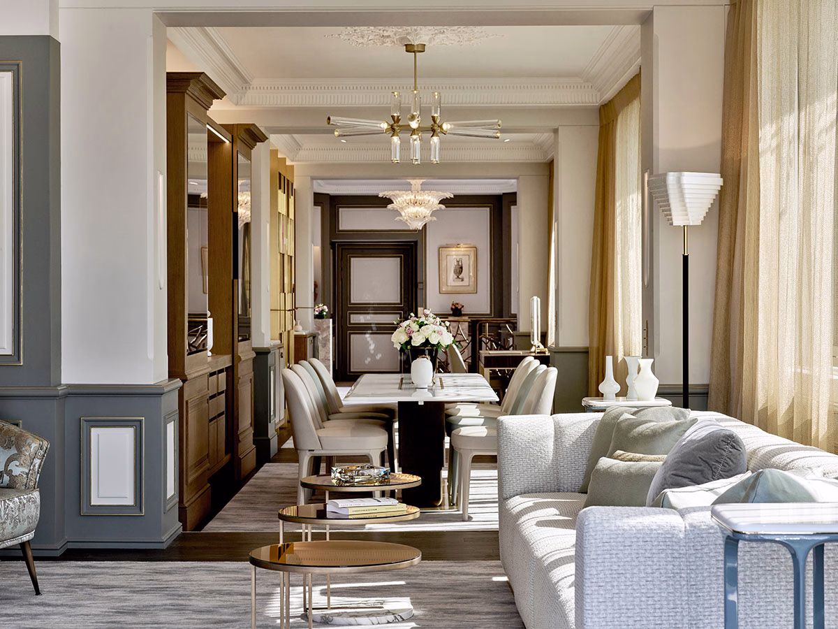 Blick durch eine luxuriöse Suite im Le Meurice, mit einem eleganten weißen Marmor-Esstisch und cremefarbenen Stühlen unter einem modernen Kristallleuchter im Hintergrund sowie einem hellgrauen Sofa mit runden Beistelltischen im Vordergrund